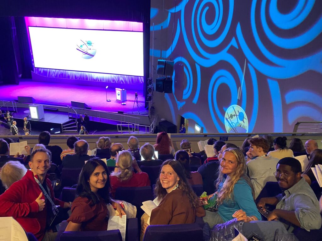 A group of people sitting in an auditorium. The stage is lit in purple and blue with the Glasgow 2024 logo projected on the wall behind them.